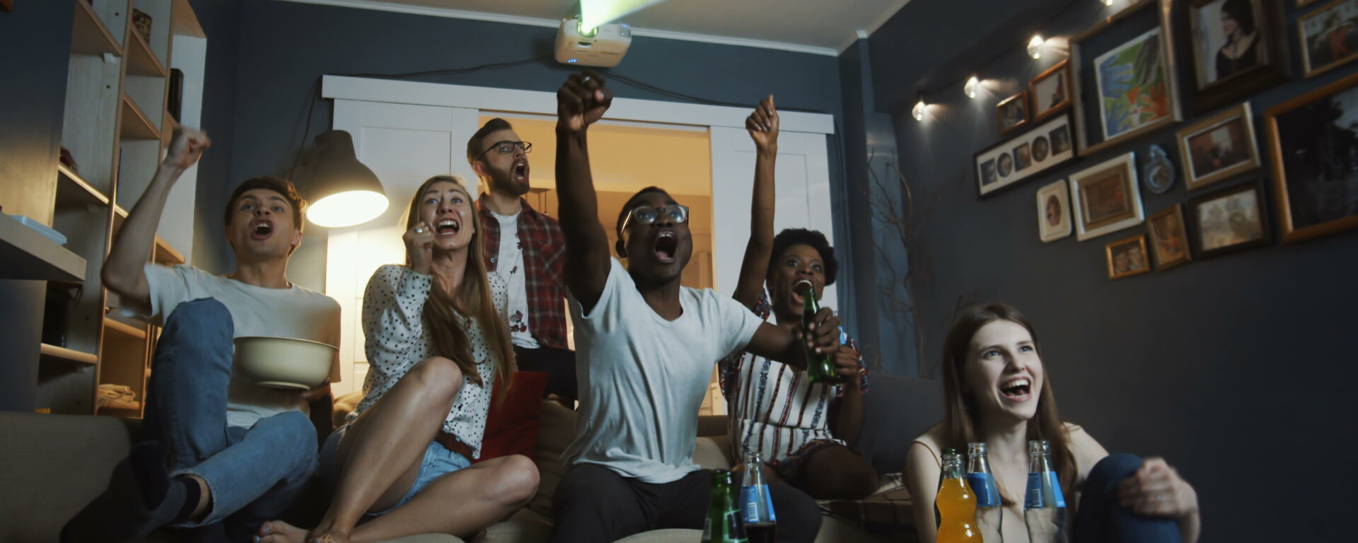 Excited young multiethnic friends get emotional and shout watching sports on a projector together at home in a home theater. Cheerful students get crazy, raising hands cheering for teams during a big football game or basketball game.
