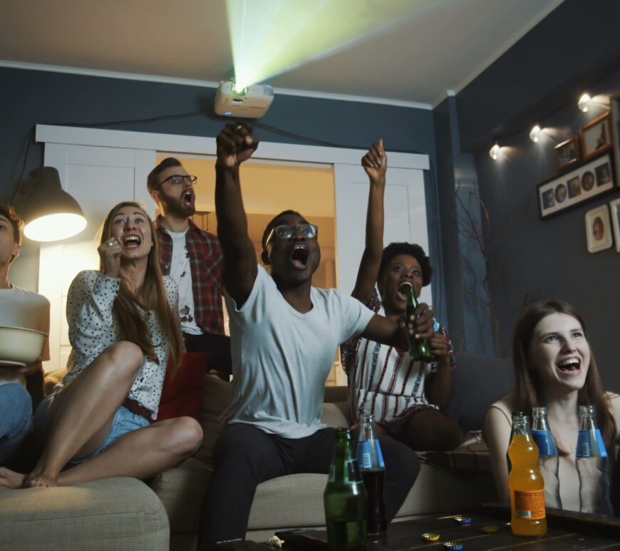 Excited young multiethnic friends get emotional and shout watching sports on a projector together at home in a home theater. Cheerful students get crazy, raising hands cheering for teams during a big football game or basketball game.