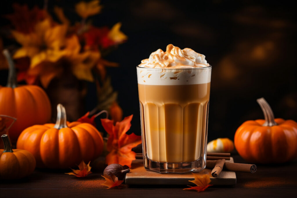 Cold brew coffee latte topped with whipped cream, pumpkin spice mix, and caramel sauce. Set next to pumpkins, orange leaves, cinnamon sticks, and nutmeg.
