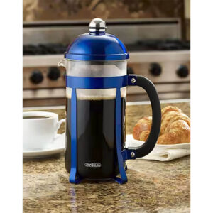 Blue french press sitting on a counter top next to a cup of coffee and a croissant.