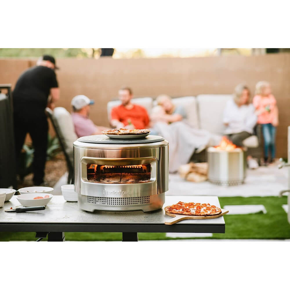 A family sitting on an outdoor couch enjoying pizza from a small portable wood-fired pizza oven.