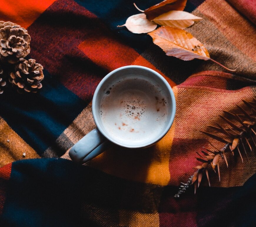 A cup of coffee on a orange and blue quilted blanket surrounded by pinecones and leaves to evoke images of fall.
