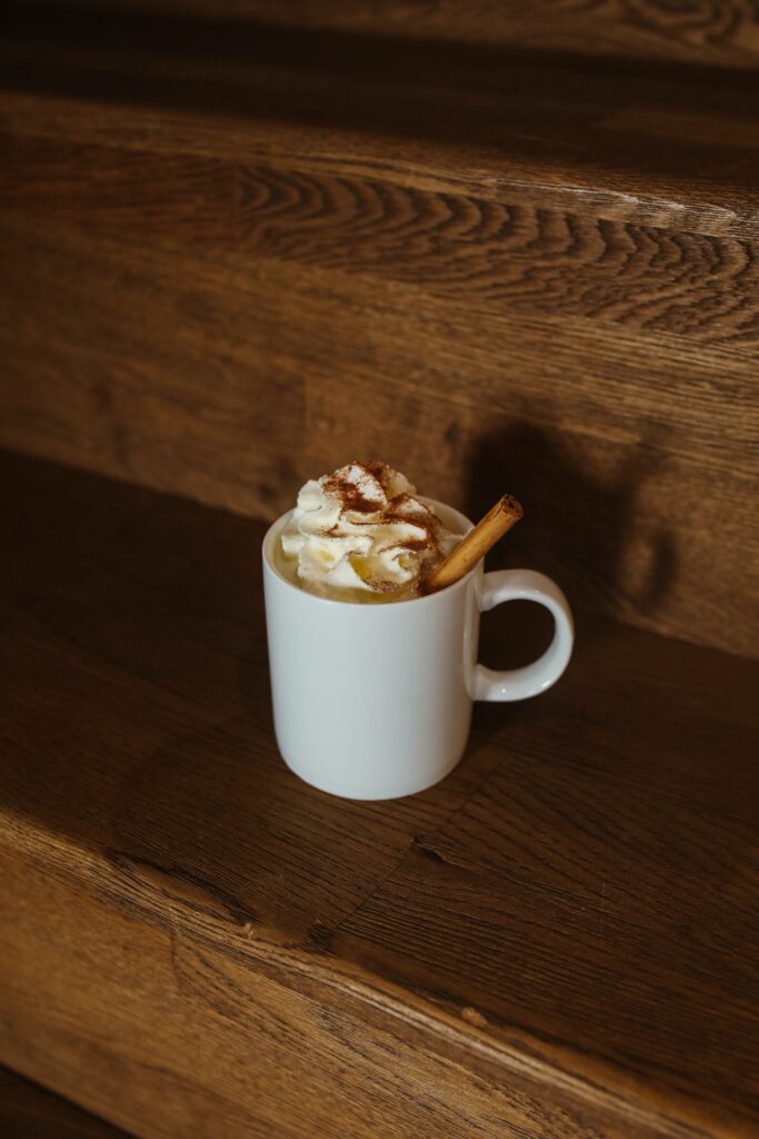White mug of coffee on a wooden bookshelf. The coffee is topped with whipped cream, cinnamon, and a cinnamon stick.