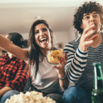 Two young couples watching sports on TV, cheering for their favorite team. They're enjoying popcorn, beer, and chips while watching the game.