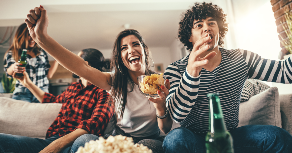 Two young couples watching sports on TV, cheering for their favorite team. They're enjoying popcorn, beer, and chips while watching the game.