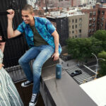 Two people using a JBL Flip 6 on a root top balcony above other apartment buildings, trees, and a street below.