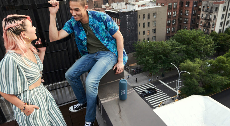 Two people using a JBL Flip 6 on a root top balcony above other apartment buildings, trees, and a street below.