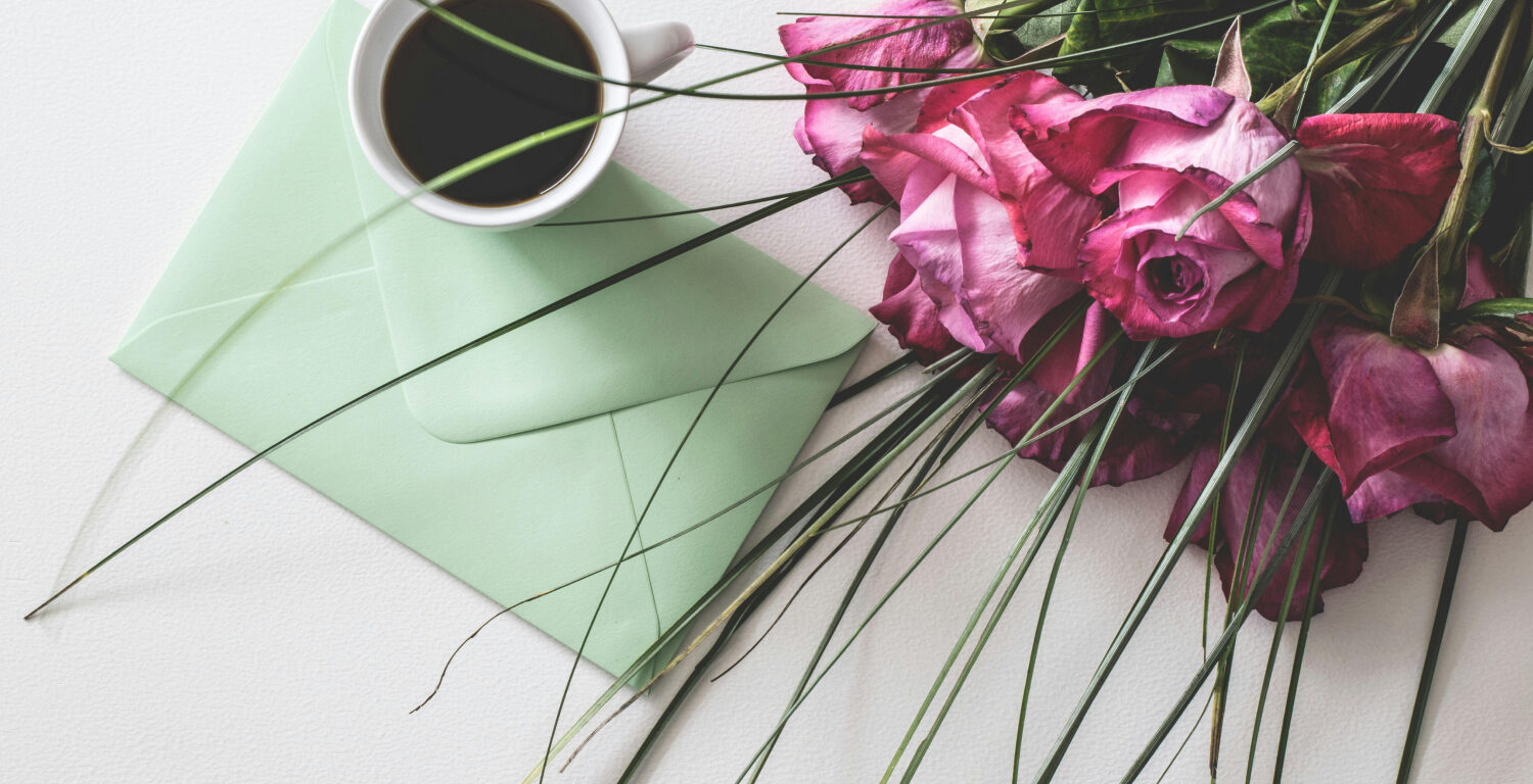 A collection of Mother's Day gifts: a cup of coffee, a bouquet of pink roses, and a card in a light green envelope.