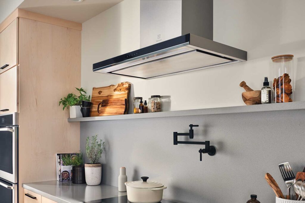 An Electrolux ventilation hood installed above an induction cooktop. A pale ceramic pot is on the cooktop. The countertops and shelf around the kitchen have cooking implements, herbs, cookbooks, and ingredients in glass jars sitting on them.