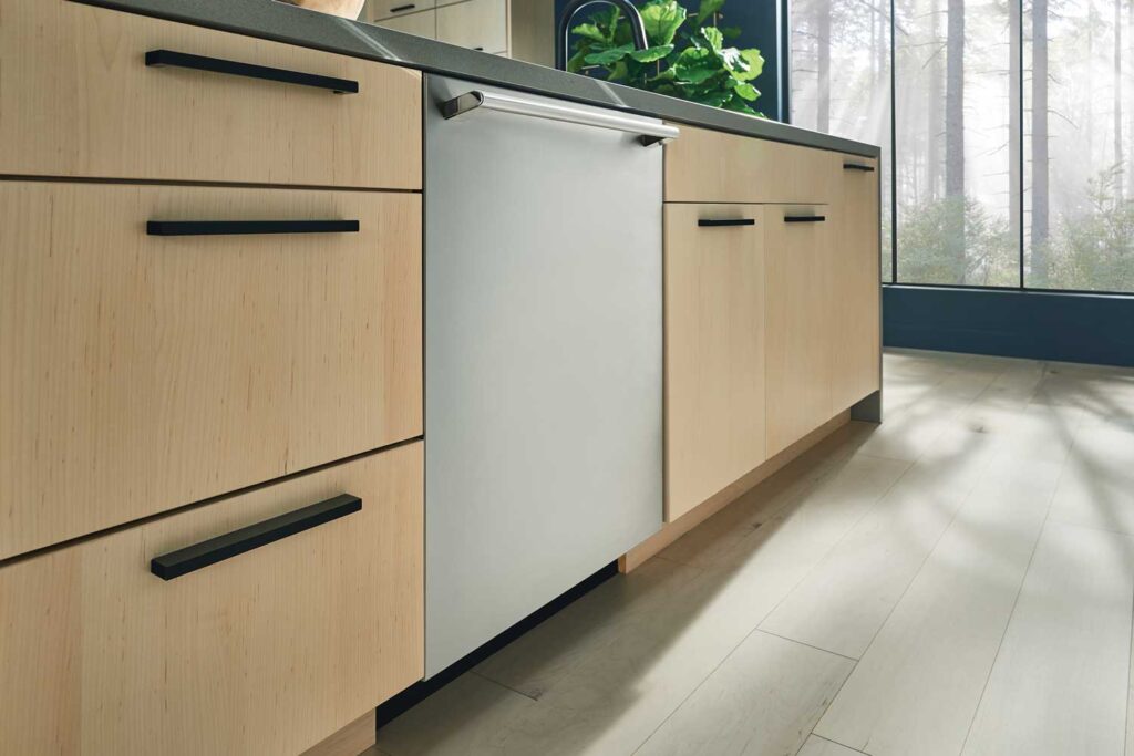 A silver Electrolux dishwasher installed under a gray countertop in a kitchen with pale tan wood drawers and cabinets. The walls are painted a dark blue, and a wide and tall window looks out into a forest, letting in natural light.