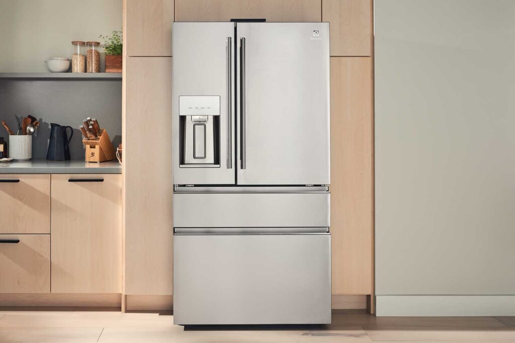 An Electrolux french door refrigerator installed in a kitchen with pale wood cabinetry. A counter extends to the left of the image where a pitcher, knife block, and cooking utensils in a holder are setting on it. 