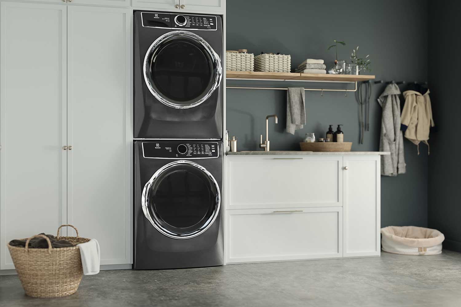 A titanium-colored Electrolux laundry center installed in pale gray laundry room cabinetry. The walls are painted dark gray, and there is a countertop next to the laundry tower with a sink and soaps sitting in a basket.