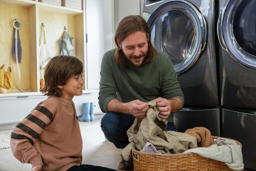 A father with shoulder-length brown hair squats next to an open Electrolux washing machine in a laundry room. He and his son are looking at their laundry that's sitting in a basket.