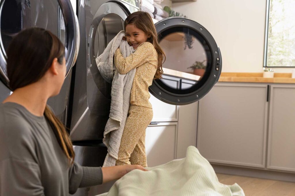 A mother looks at her daughter who is taking a warm blanket out of an Electrolux dryer that just finished a cycle. The mother is folding blankets while sitting on the laundry room floor.